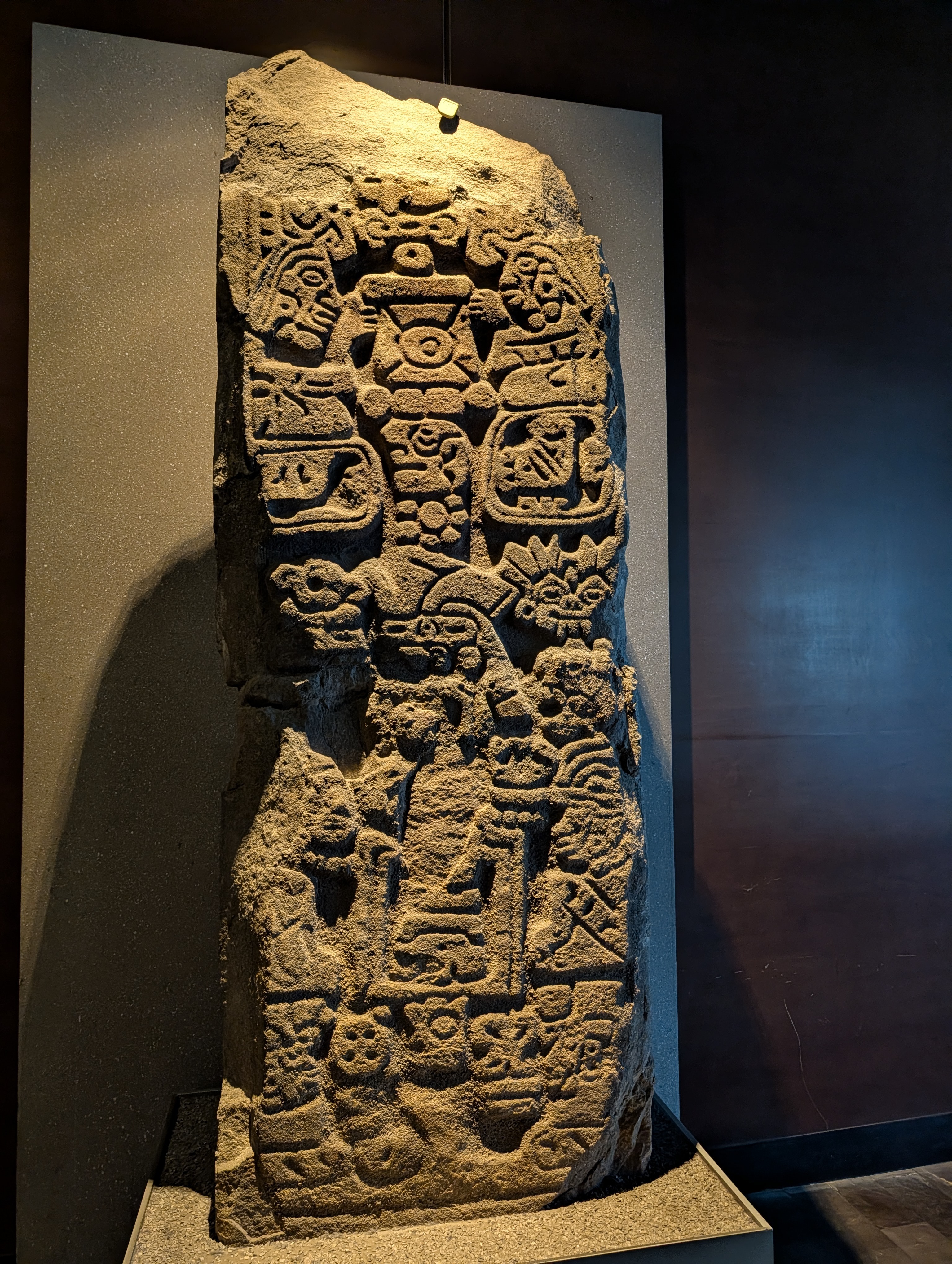 A carved stone stela — a freestanding monument slab covered top to bottom in relief carving, densely packed with glyphic cartouches (the boxed symbols) alongside a larger central figure in elaborate headdress and regalia.