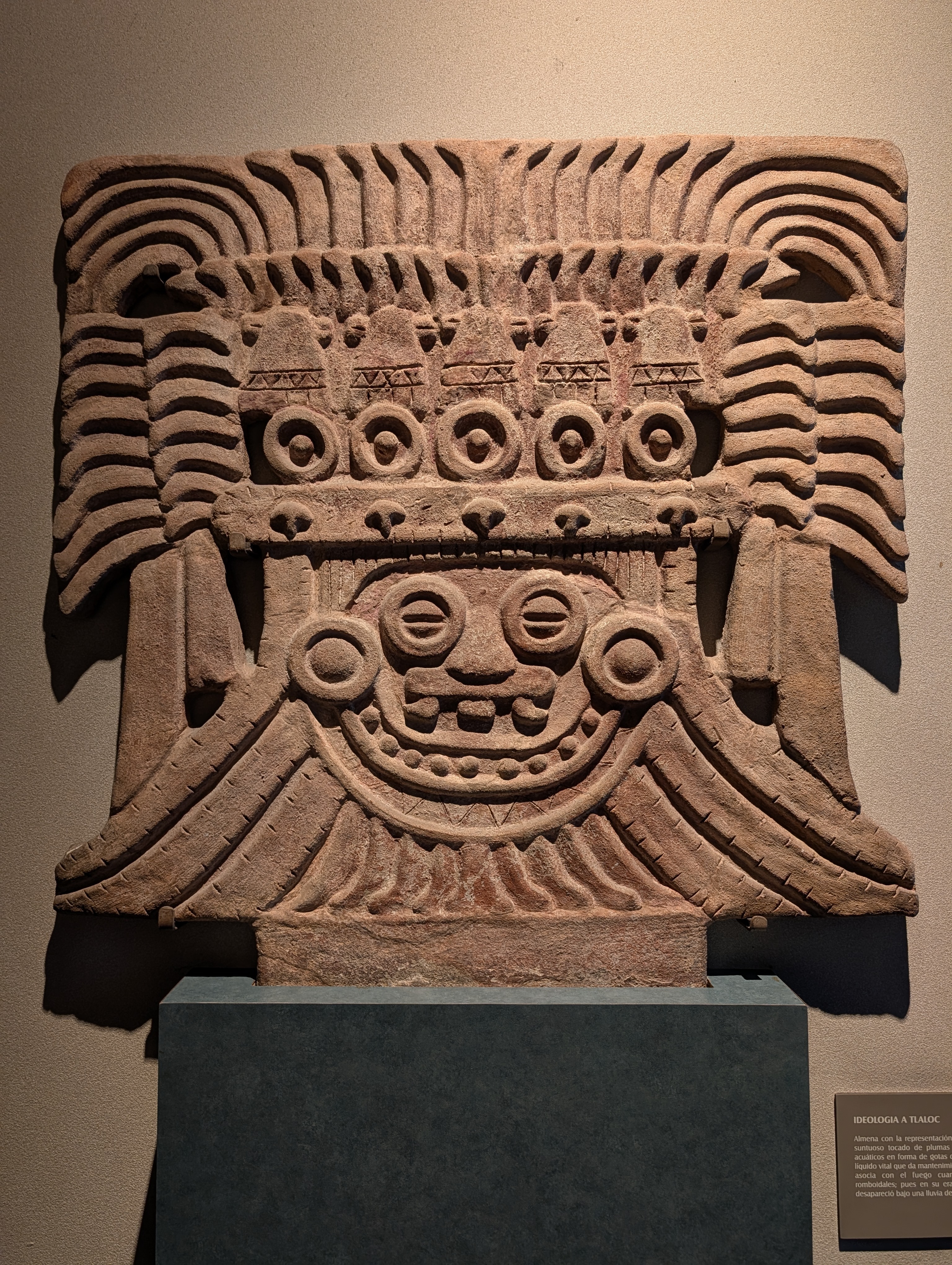 Ideología a Tlaloc.  A carved almena depicting Tlaloc, the rain god, identifiable by his characteristic goggle-eyed face, fanged mouth, and the elaborate feathered headdress with the row of circular water-drop or year-sign motifs across the brow. The rippling, feather-like carved bands framing the face represent his sumptuous plumed headdress, tied to water and fertility imagery.
