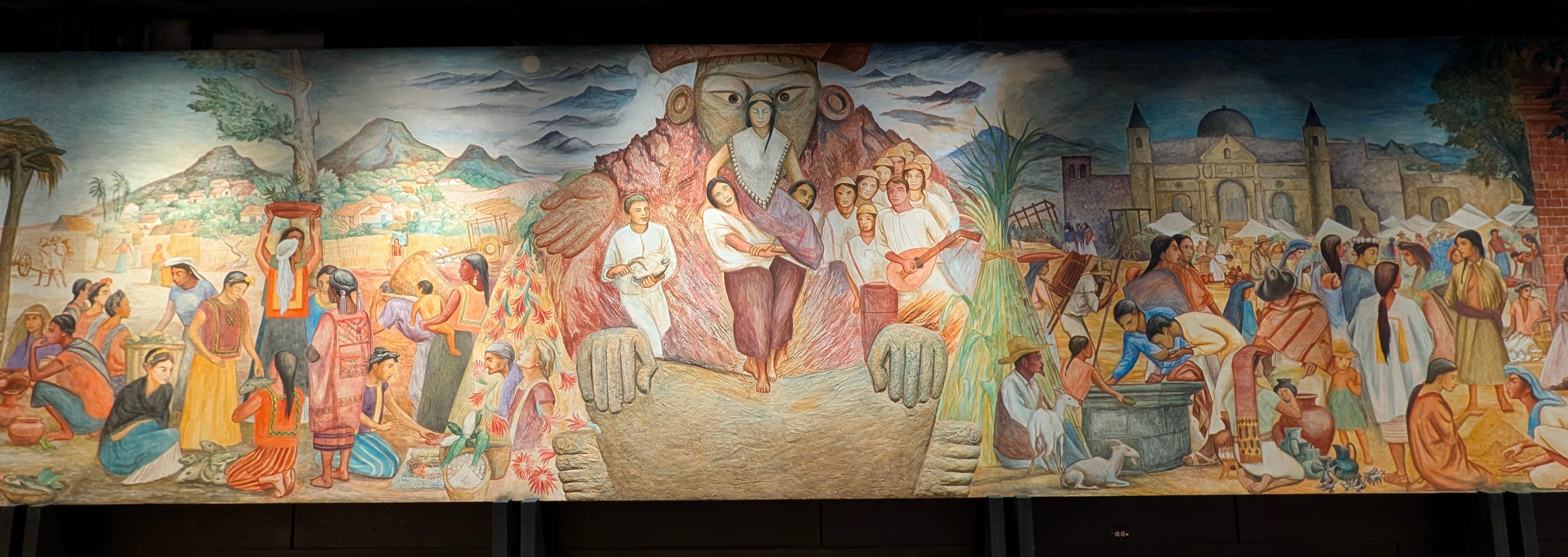 This is a large-scale mural depicting rural/village life in Mexico, framed by a giant sculpted figure emerging from a mountain (likely a stylized volcano/mountain deity, possibly evoking Coatlicue or a mountain-earth spirit) at center.